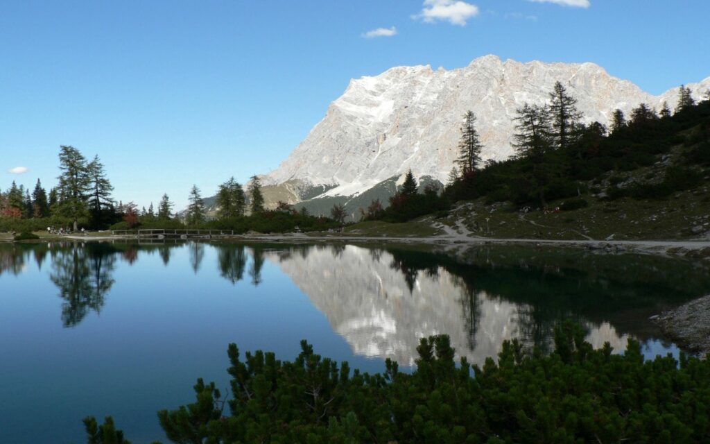 Serene mountain landscape reflected in calm lake with lush trees rugged rocks under clear sky Wallpaper