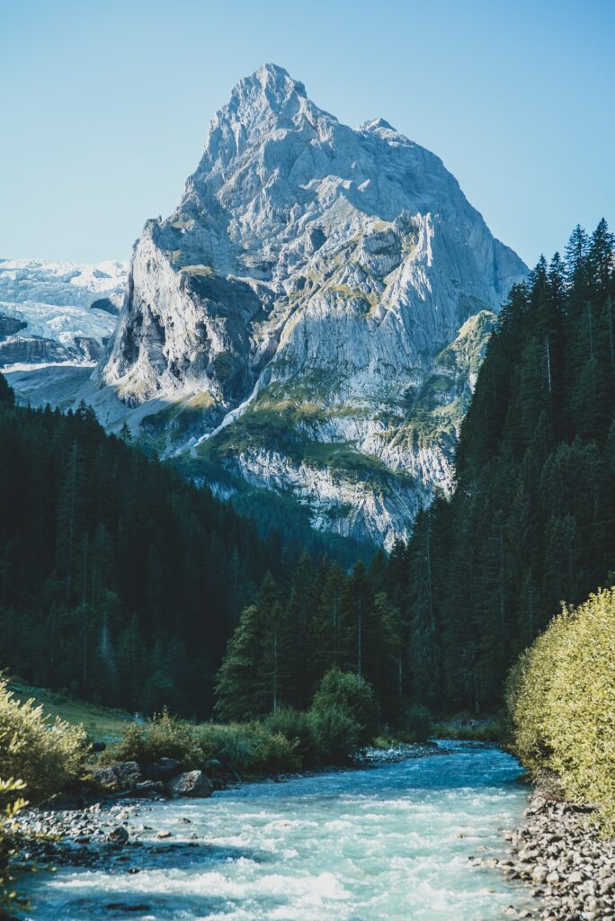 Crystal clear river winding through lush green forest with towering mountain under blue sky wallpaper