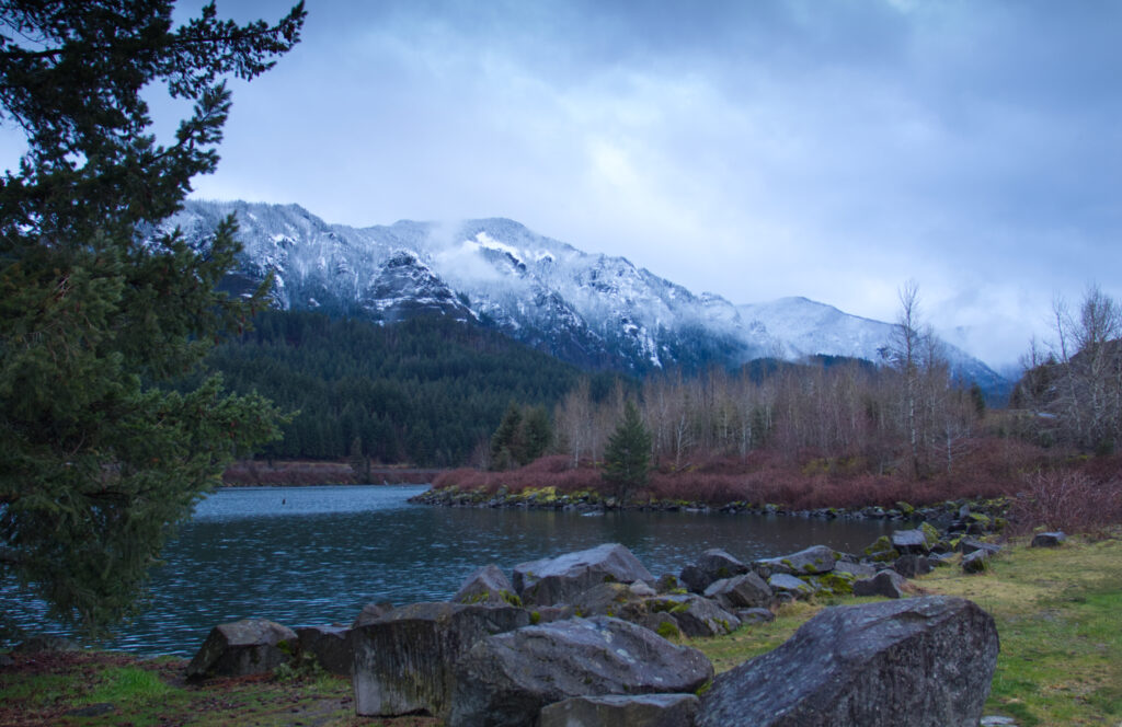 Serene mountain landscape with snow peaks calm lake reflective cloudy sky rugged shore stones Wallpaper