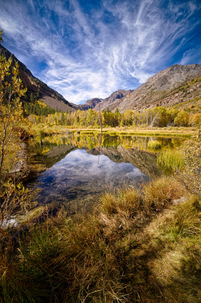 Serene mountain landscape with clear pond reflecting autumn foliage and rugged peaks under blue sky Wallpaper