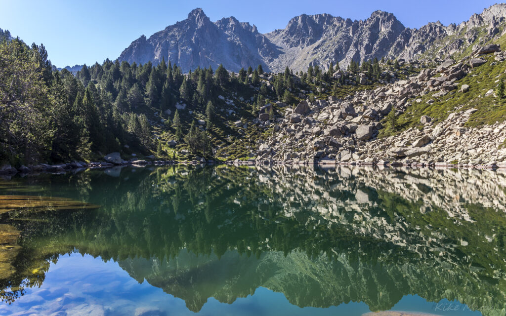 Tranquil lake reflecting rugged mountains and lush pine trees in vibrant blues and greens Wallpaper