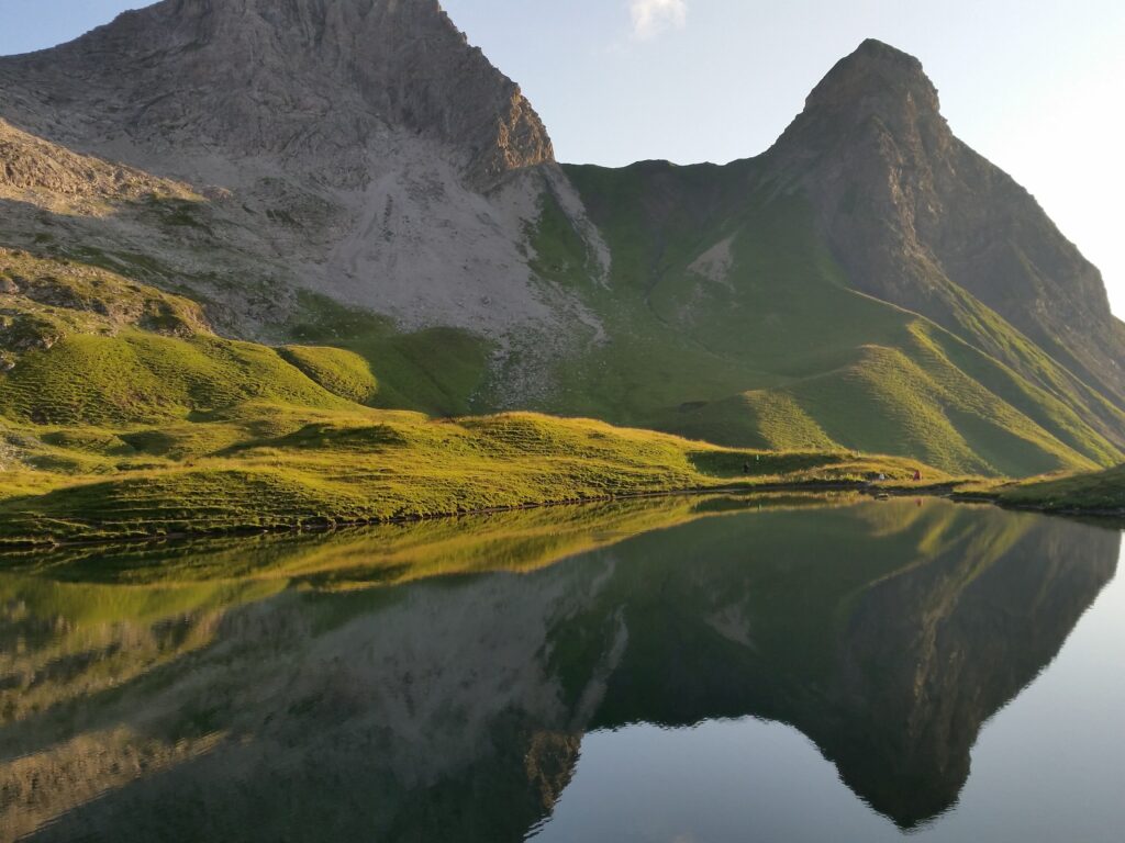 Serene mountain landscape with rocky peaks lush grass and tranquil lake reflecting soft morning light Wallpaper