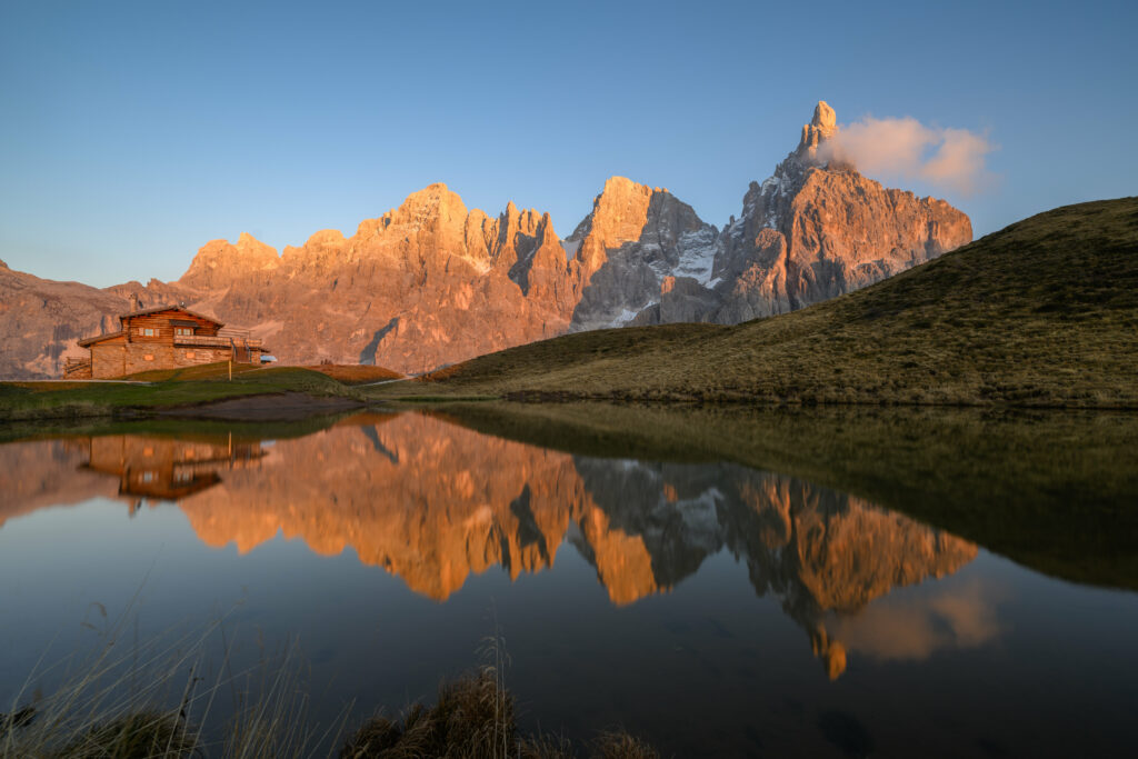 Serene mountain landscape with rugged rocks cozy cabin green hillside reflected in calm lake Wallpaper