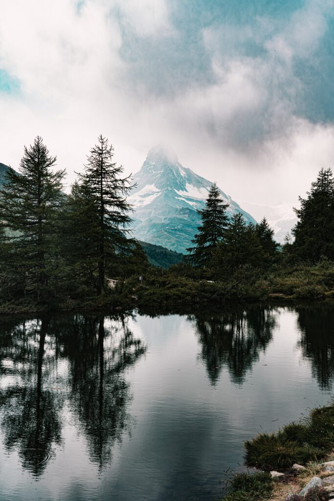 Serene mountain landscape with spruce trees and lake reflecting snow-capped peak and clouds Wallpaper