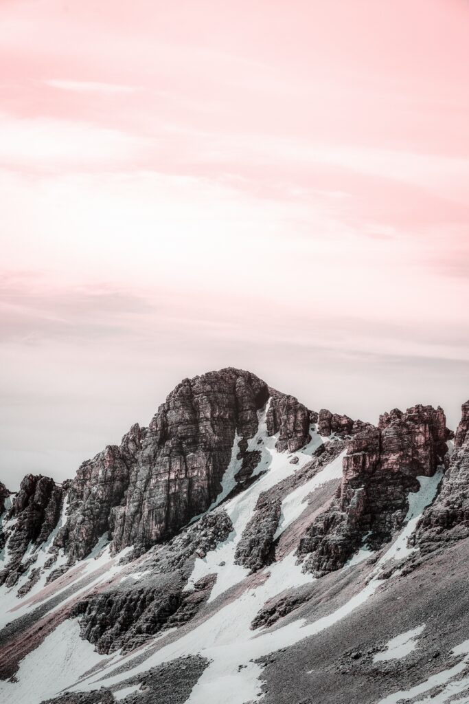 Mountain peak with rugged stones and snow under a soft pink and white serene sky wallpaper