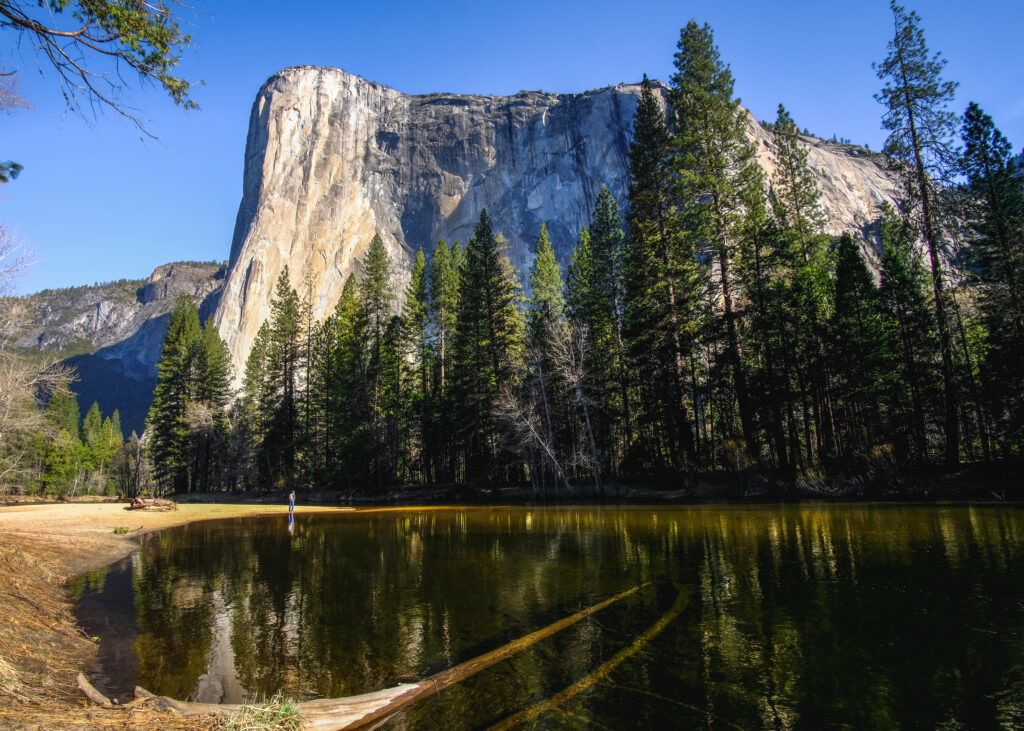 Dramatic rock formation above pine forest reflected in calm lake under clear blue sky wallpaper
