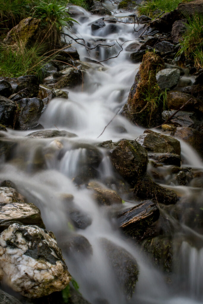 Serene mountain stream flowing over mossy stones with smooth misty water and green grass background Wallpaper