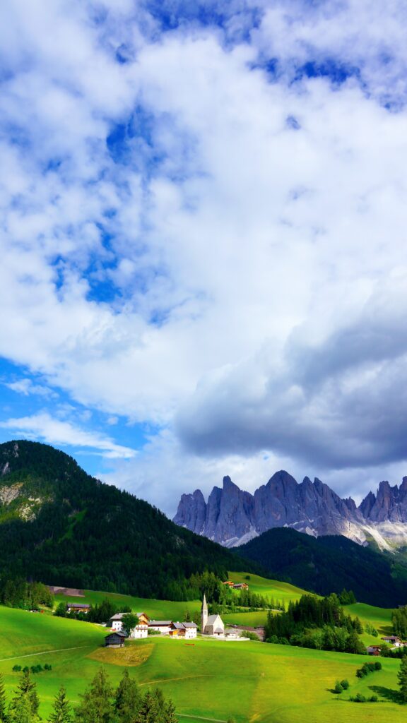 Vibrant mountain range wallpaper with green grass village trees and dramatic cloudy blue sky background