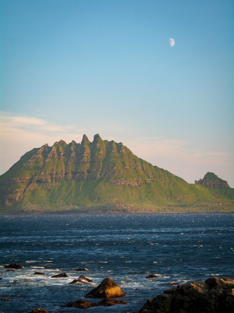Lush green mountain over calm sea with rocky shore under clear blue sky and distant moon wallpaper