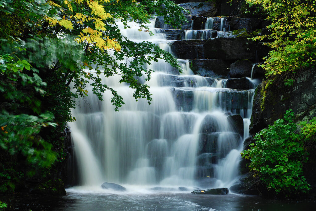 Serene multi-tiered waterfall over dark rocks with lush green and yellow trees misty water flow Wallpaper