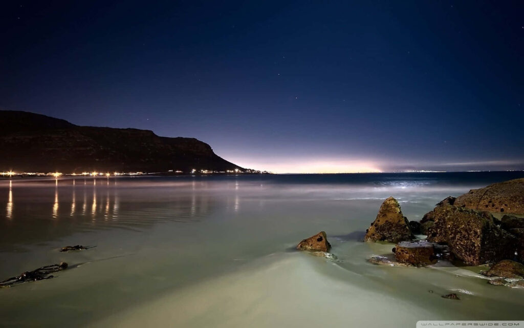 Tranquil Night Beach: Reflective Waters & City Lights Under Stars Wallpaper