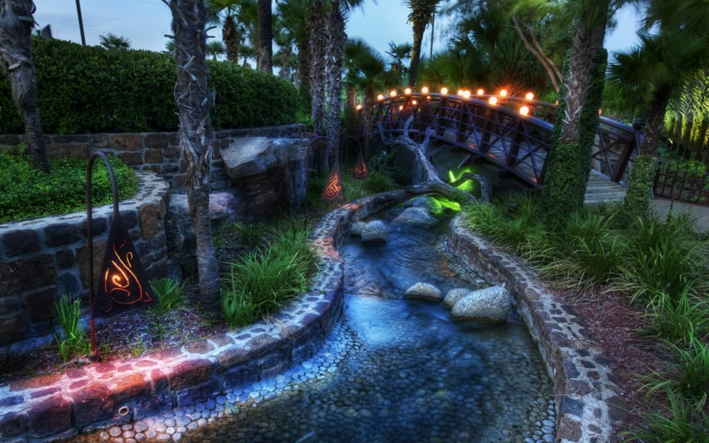 Tranquil night garden with illuminated bridge over stream lush greenery smooth stones wallpaper