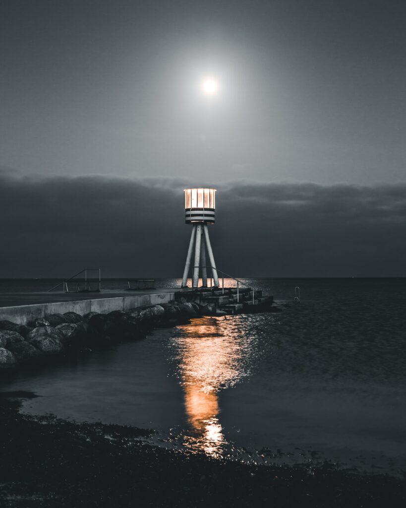 Modern lighthouse glowing on pier at night with calm sea stones moonlight serene wallpaper background