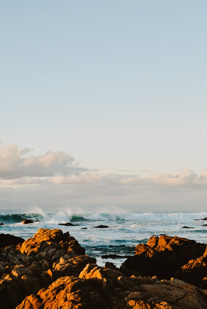 Sunlit ocean waves crashing on rugged rocks with soft blue water and warm golden tones Wallpaper