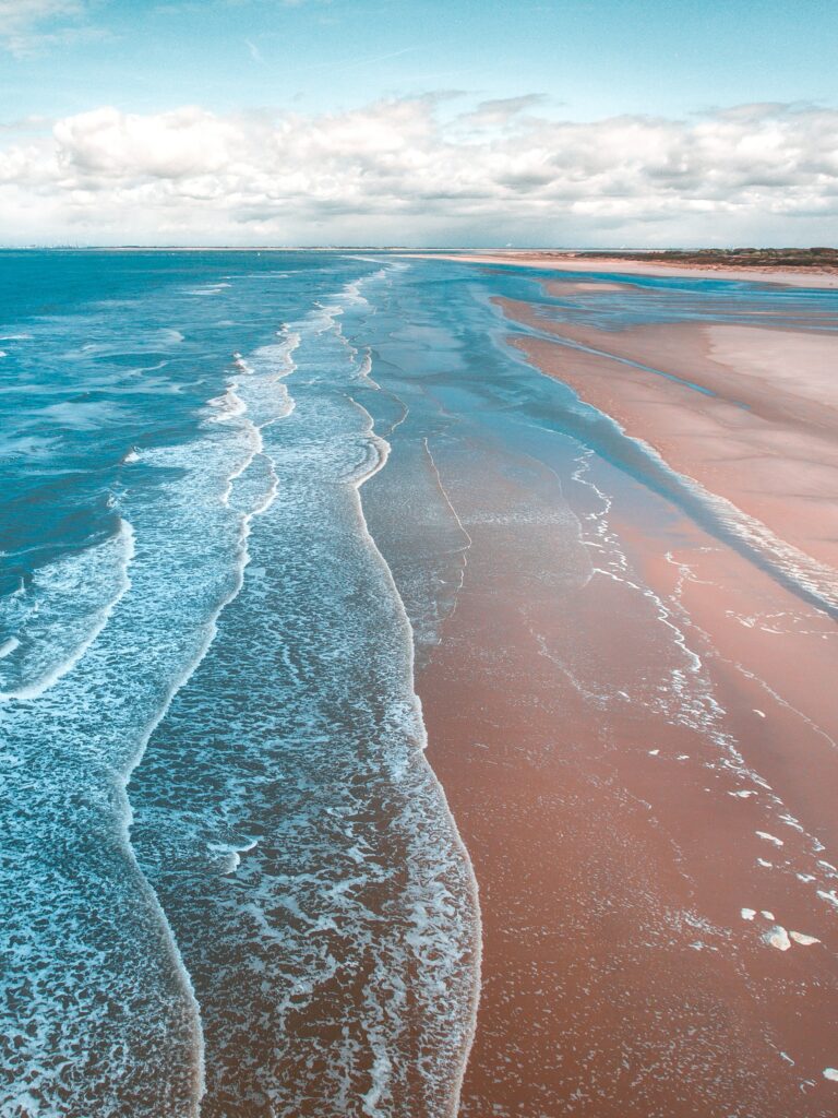 Aerial view of ocean waves gently rolling onto sandy shore with white foam and clear blue water Wallpaper