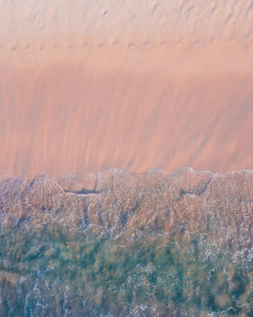 Aerial view of pastel blue pink waves gently washing on sandy coast serene wallpaper background