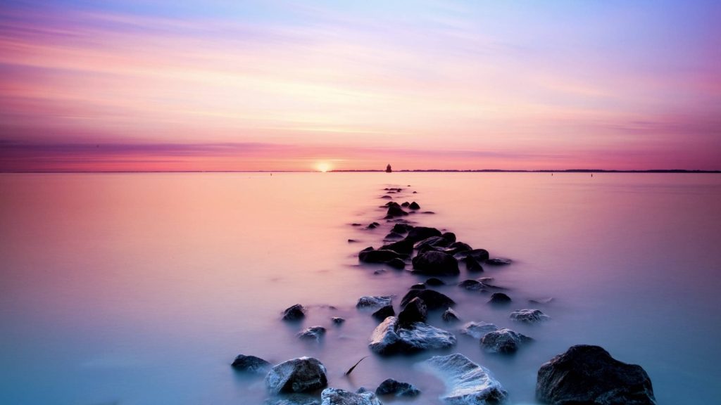Soft pink clouds sunset over calm waters with rocks leading horizon serene wallpaper background