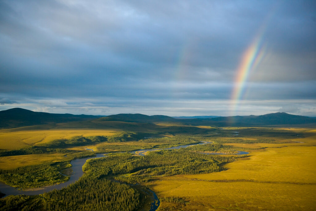 Vibrant rainbow over green field and winding river with cloudy sky and golden light landscape Wallpaper