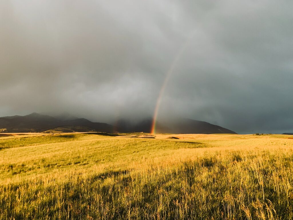 Vibrant rainbow over sunlit grassy field with mist fog dramatic clouds and dark mountains background Wallpaper