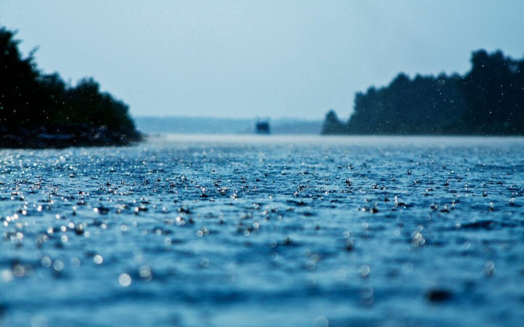 Raindrops creating ripples on water with sparkling reflections and blurred blue shoreline background Wallpaper