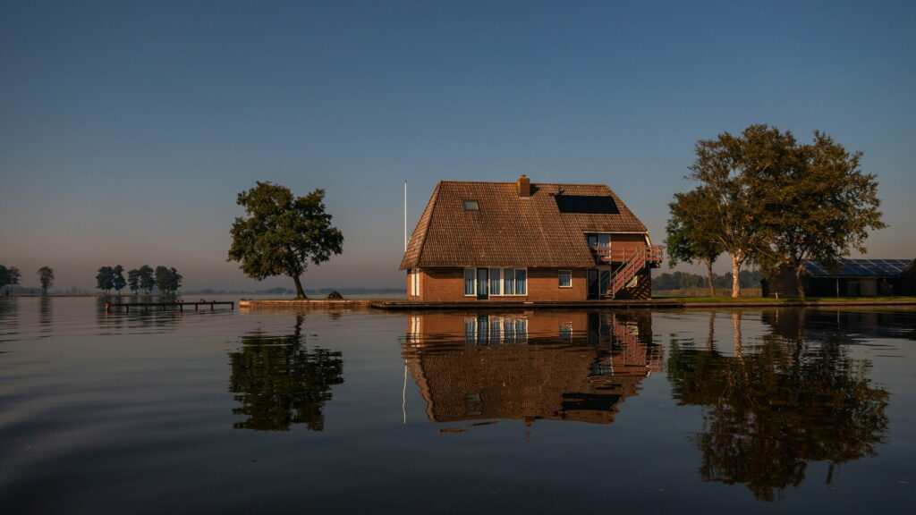 Serene Reflection: Tranquil Waters Reflecting a House and Tree under a Clear Blue Sky Wallpaper