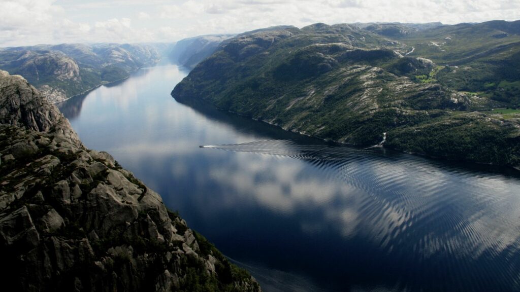 Boat gliding on calm river with rugged rocks and green coasts under partly cloudy sky wallpaper