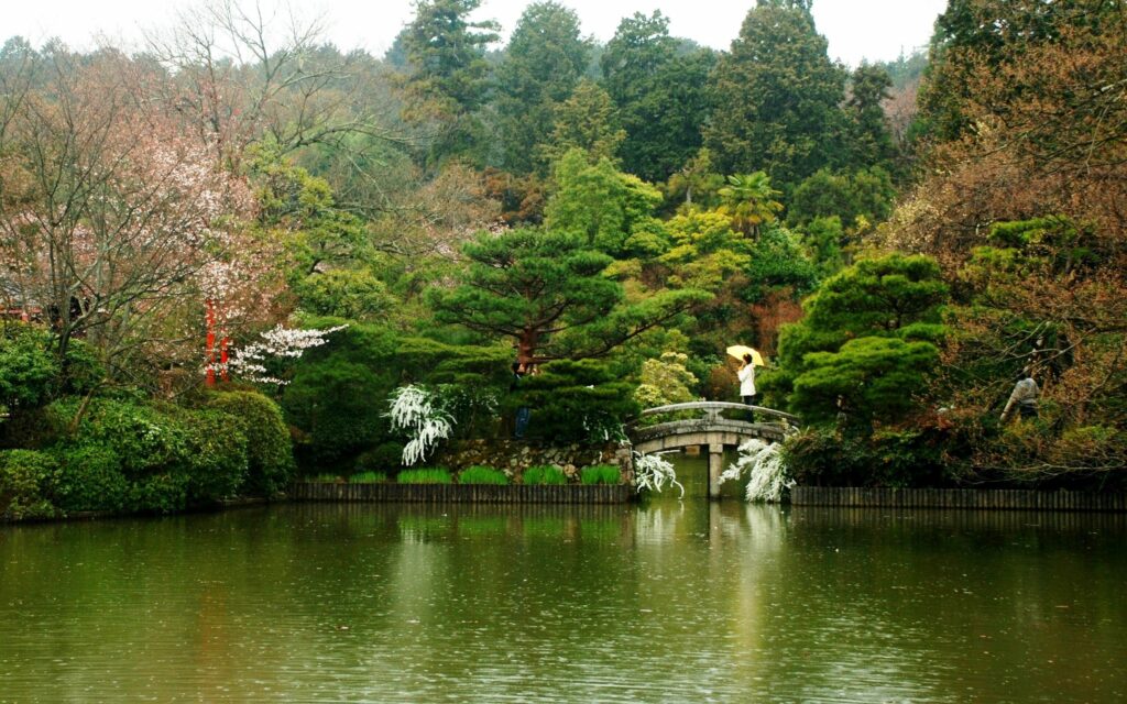 Serene river flowing under stone bridge with lush trees and person holding umbrella crossing Wallpaper