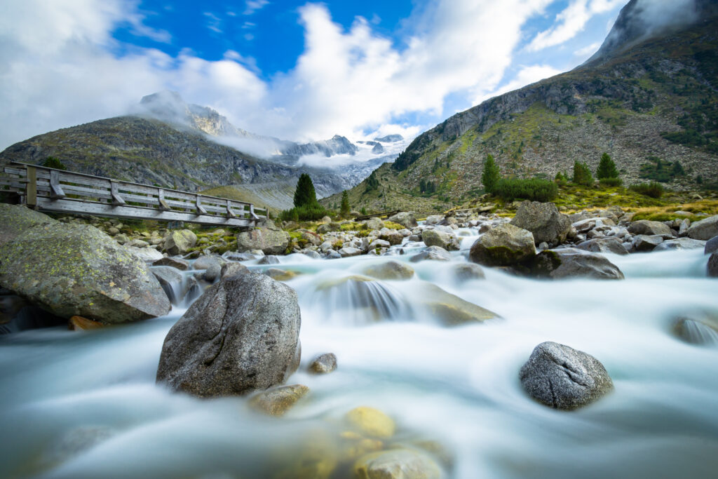 Serene river flowing over smooth stones with lush green mountains and blue sky background Wallpaper