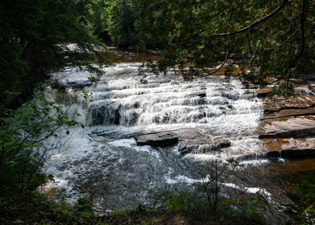 Serene river cascading over stone steps with lush green forest and sunlit flowing water wallpaper