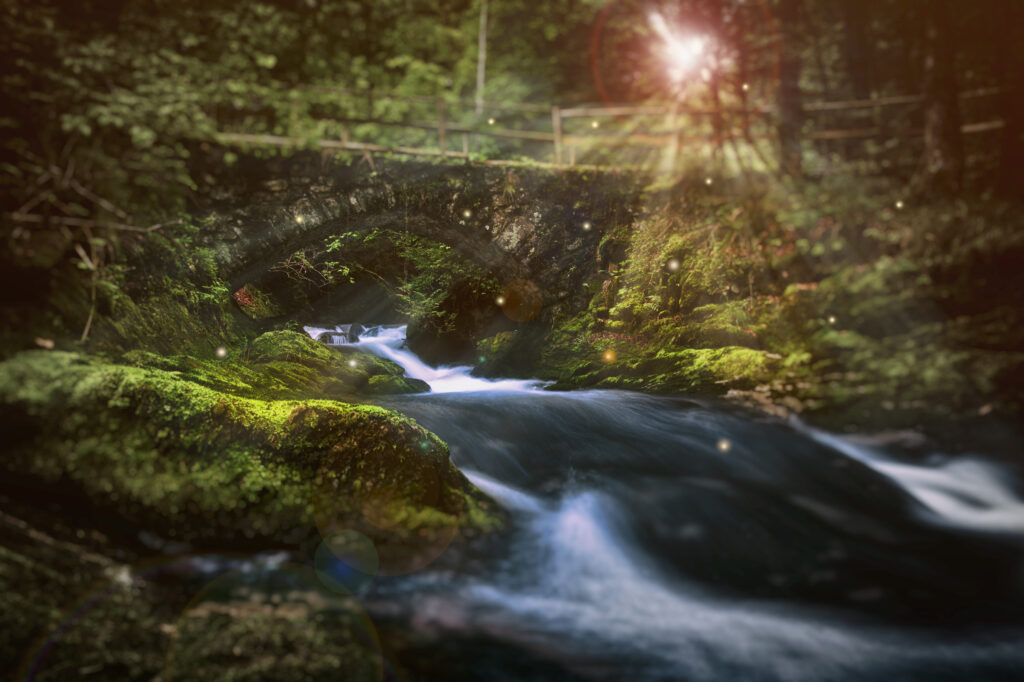 Serene river flowing under old stone bridge with lush greenery and sunlight streaming through trees Wallpaper