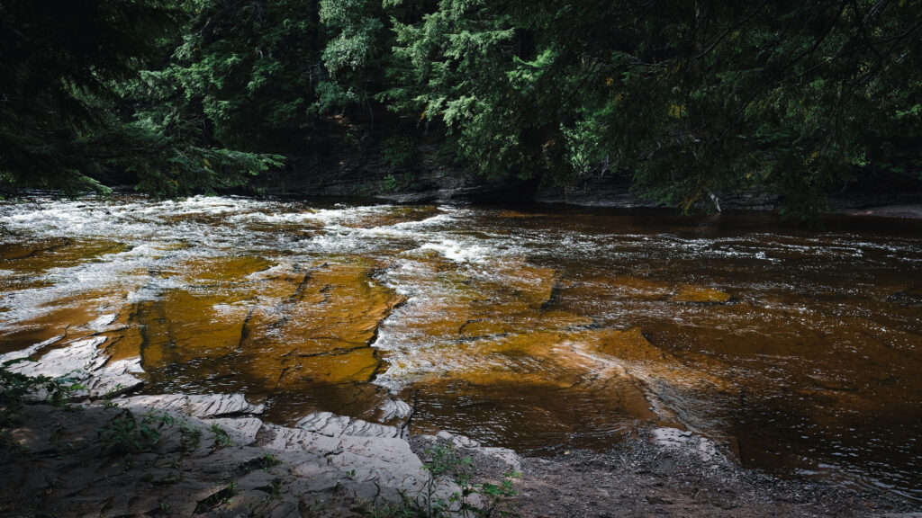 Tranquil river flowing through dense green forest with weathered stones and sunlight rays Wallpaper