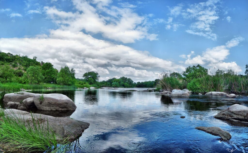 Serene river with smooth stones lush greenery clear reflective water and vibrant blue sky wallpaper