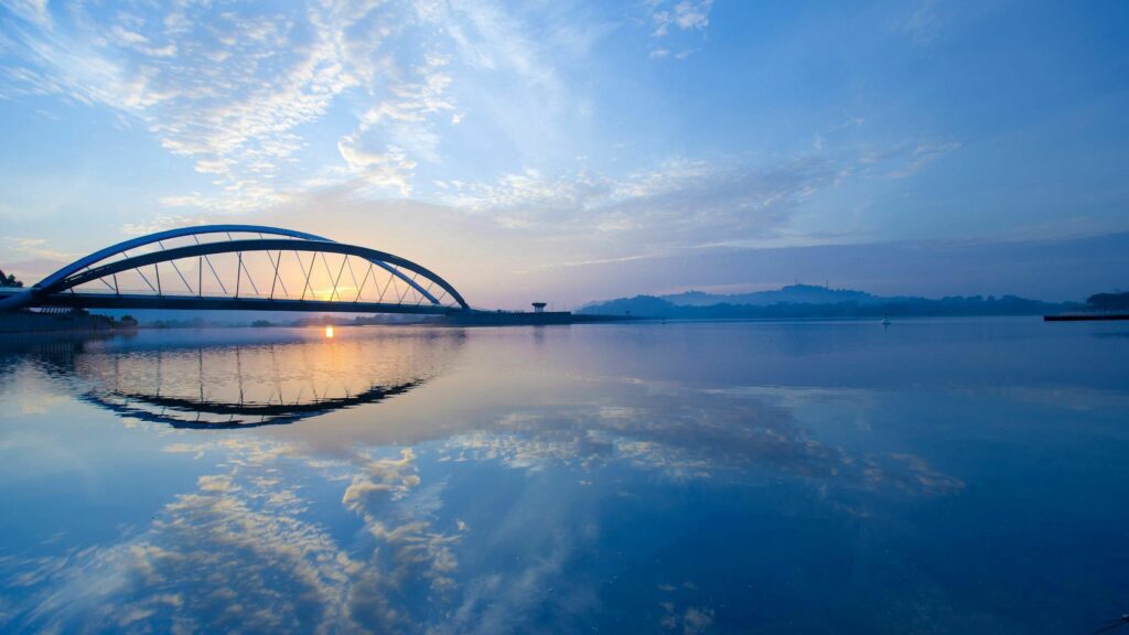 Serene river with modern arched bridge reflecting golden sunset sky and soft blue hues Wallpaper