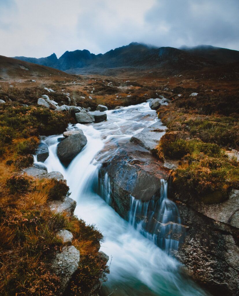 Serene river waterfall over smooth stones with rugged mountains under cloudy sky wallpaper background