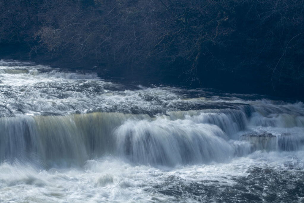Serene river with cascading water over smooth rocks soft blue white tones and leafless branches background Wallpaper