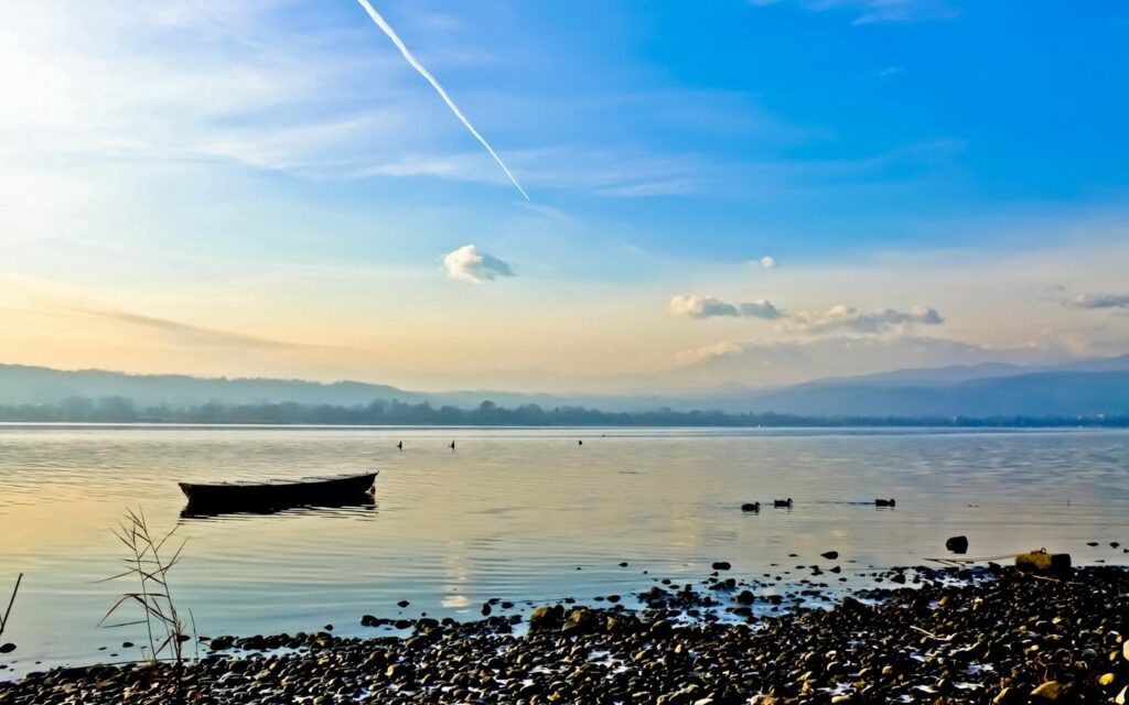 Solitary boat on calm river near smooth stones with distant hills and blue sky background Wallpaper