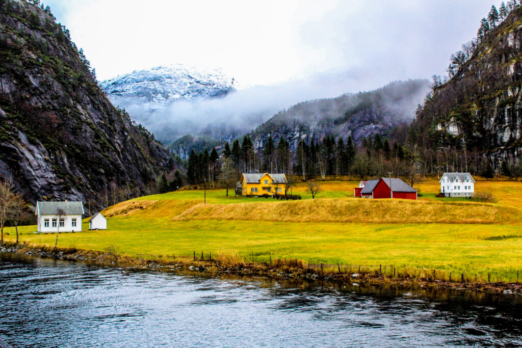 Peaceful riverside houses on grassy shore with misty mountains dense forests vibrant colors wallpaper