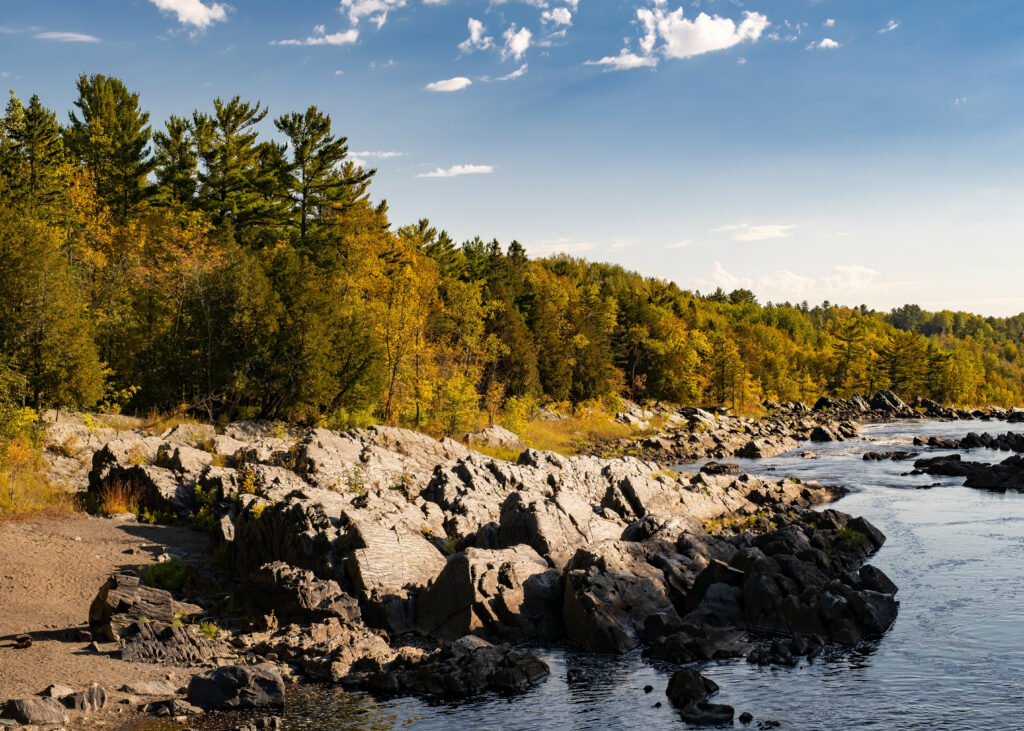 Serene riverside landscape with smooth stones lush green and golden trees reflecting blue sky Wallpaper