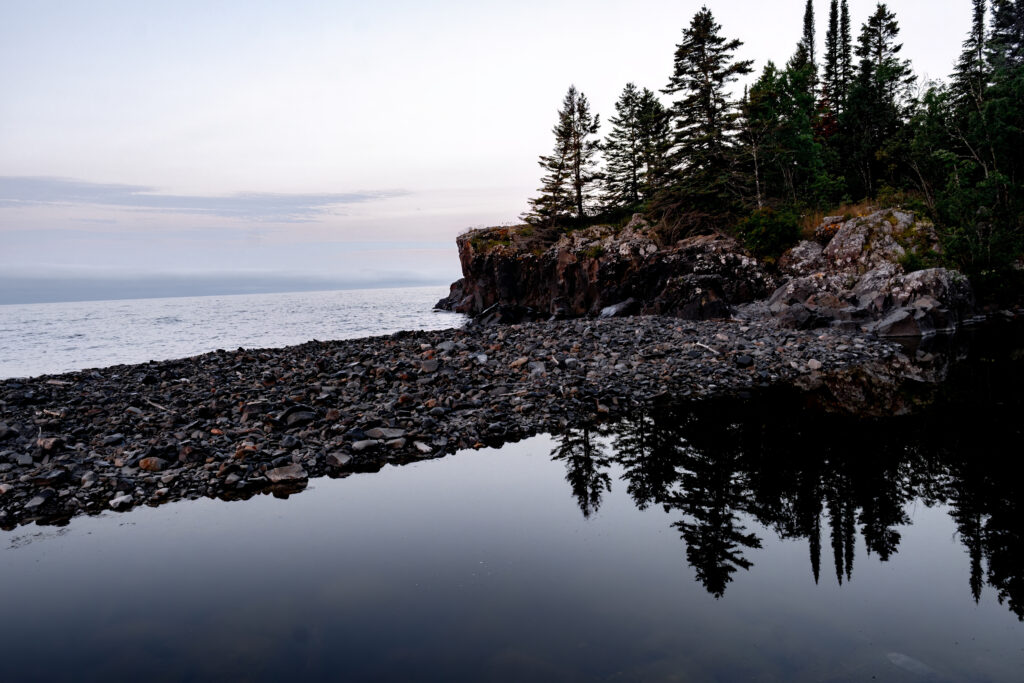 Tranquil rugged coastline with dark pebbles rocky cliffs dense trees calm water reflecting sky Wallpaper
