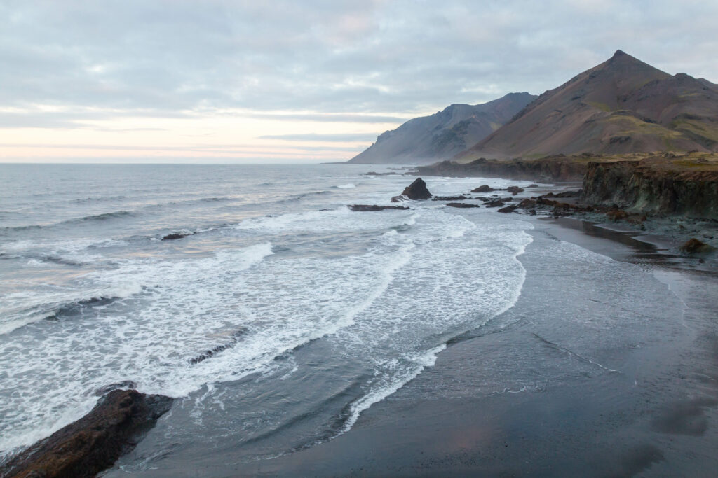 Rugged mountains meet dramatic coastline with waves on dark sand under cloudy sky wallpaper background
