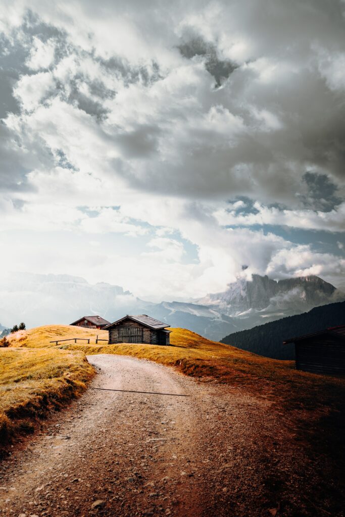 Winding dirt road past rustic wooden houses on golden hills with mountains and dramatic cloud skies Wallpaper