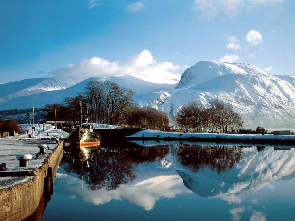 Snow-capped Scottish mountains reflected in calm lake with boat dock bridge and bare trees winter Wallpaper