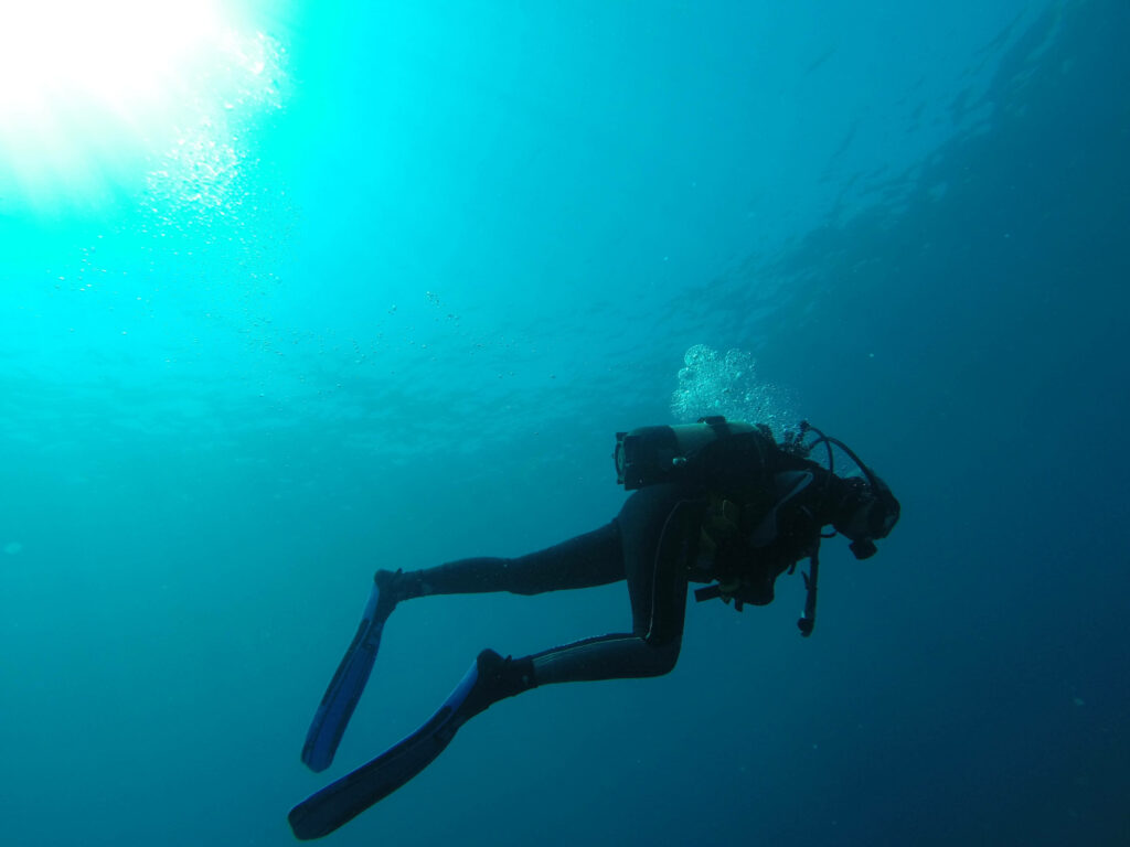 Scuba diver swimming in deep blue water with sunlight rays bubbles fins and air tank visible Wallpaper