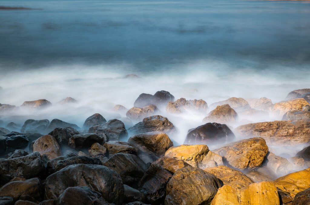 Sea mist swirls around rugged stones with blue and gold tones in a tranquil foggy coastal scene Wallpaper