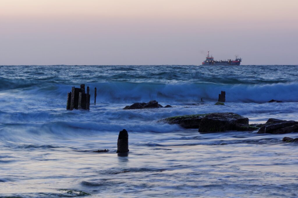 Serene dusk seascape with waves on rocks old wooden pilings and distant ship silhouette wallpaper