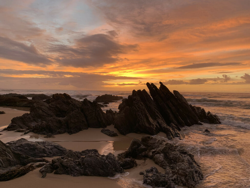 Jagged rocks on sandy shore with gentle waves under vibrant orange sunset serene seaside wallpaper