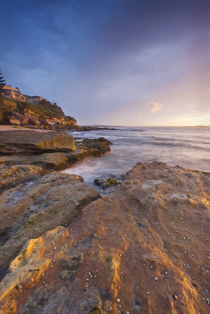 Serene seaside sunset with rugged rocks gentle waves golden light and coastal cliff houses wallpaper