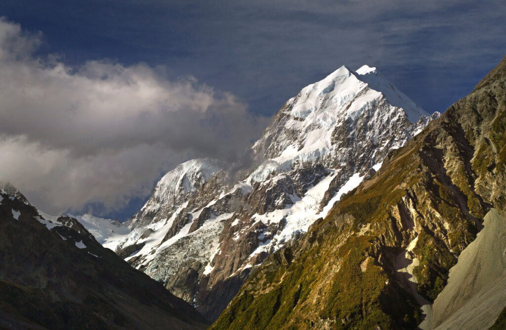 Snow-capped mountain peaks with lush rugged terrain and wispy clouds under a deep blue sky Wallpaper