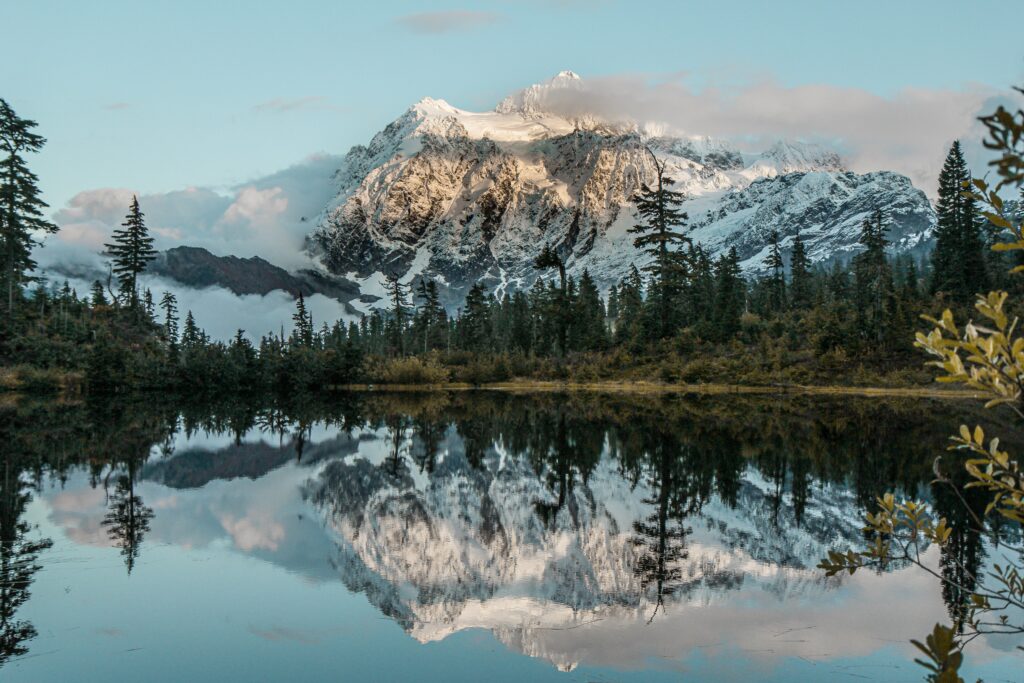 Snow-capped mountains reflected in calm lake with lush trees and misty clouds serene landscape Wallpaper