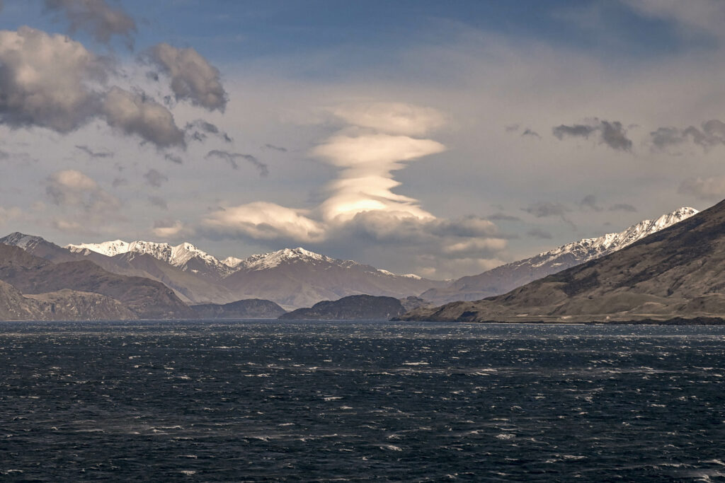Snow-capped rugged mountains over deep blue sea with gentle waves and swirling dramatic clouds Wallpaper